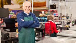Senior male standing, with arms crossed, in front of cash register,